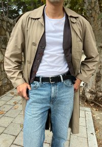 Beiger Trenchcoat mit geknöpftem Front und großen Revers, getragen über einem weißen T-Shirt, kombiniert mit hellblauen Jeans und einem schwarzen Gürtel.