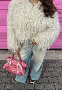 Bílá chlupatá bunda, vybledlé světle modré džíny s širokými nohavicemi a růžová kabelka s řetízkovými detaily, držená v rukou ozdobených několika prsteny.