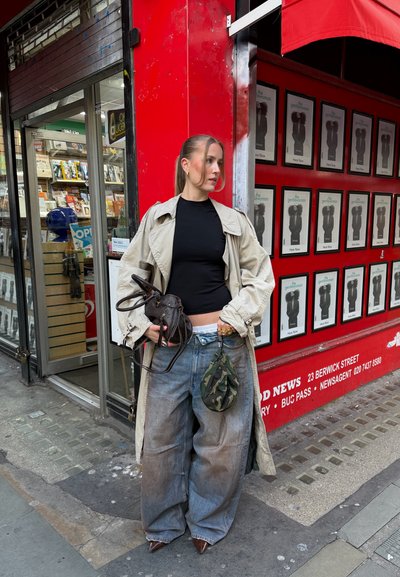 Mujer con abrigo beige, top negro, vaqueros anchos, sosteniendo dos bolsos, de pie junto a una tienda roja con carteles enmarcados y detalles del quiosco de prensa.