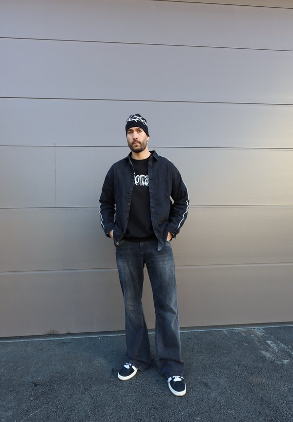 Veste en jean noir avec des rayures blanches sur les côtés, sweat-shirt noir avec du texte, pantalon en jean foncé, et baskets noires avec semelles blanches.