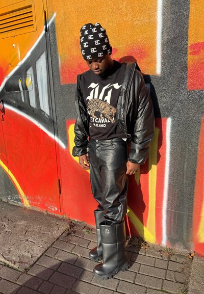 Chaqueta acolchada negra, camiseta gráfica negra con estampado de leopardo, pantalones de cuero negro brillantes y botas de goma negras hasta la rodilla.