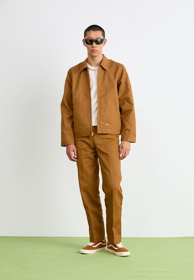 Conjunto de chaqueta y pantalones de algodón marrón con un corte relajado, combinado con una camiseta blanca y zapatillas marrones. El modelo lleva gafas de sol oscuras.
