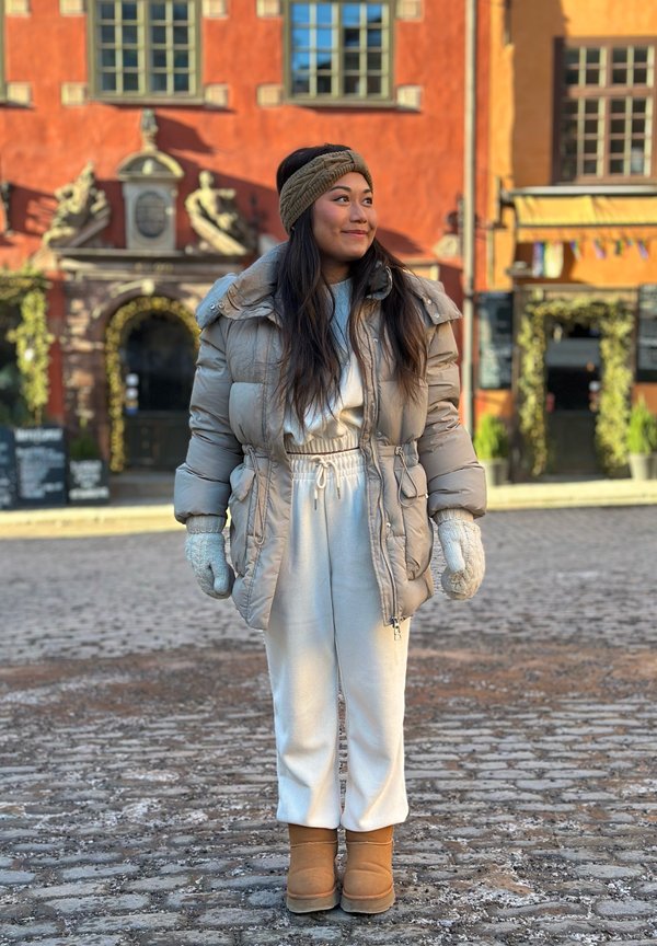 Junge Frau in beiger Winterjacke, weißen Jogginghosen, Fäustlingen, Stirnband und Stiefeln steht auf einer Kopfsteinpflasterstraße vor einem bunten Gebäude.