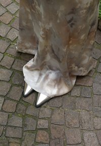 Ασημένιες γόβες με μυτερή μύτη και γυαλιστερή επιφάνεια, συνδυασμένες με ένα ρέον φόρεμα tie-dye σε απαλά μπεζ και καφέ χρώματα, που ακουμπά σε λιθόστρωτο.