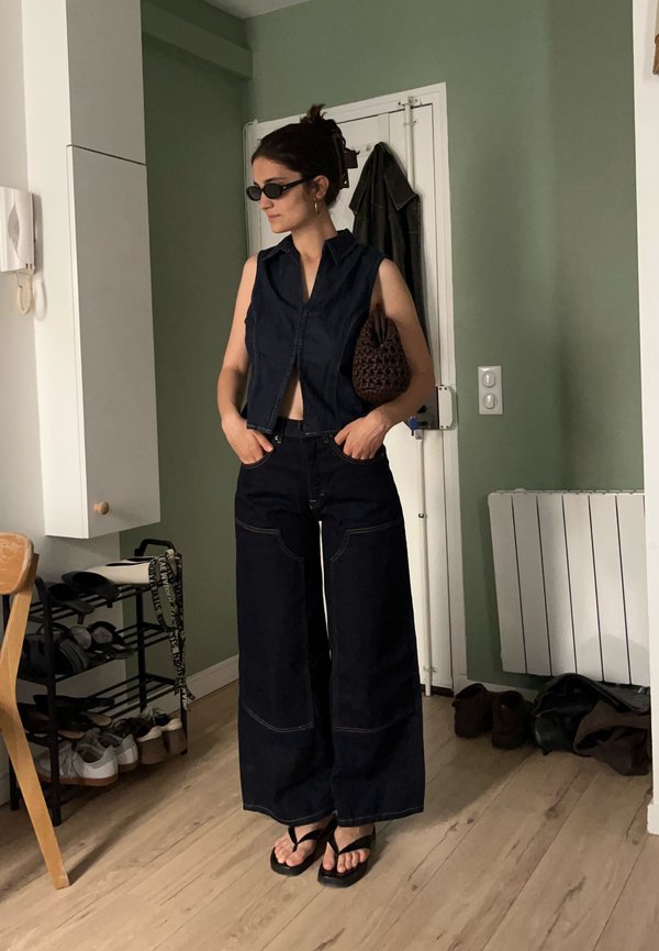 Žena so zelenou tmavou vesta bez rukávov, široké čierne nohavice, čierne sandále, slnečné okuliare a drží hnedú textúrovanú tašku v interiéri pri dverách.