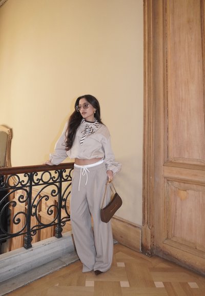 Mujer vestida con crop top beige y pantalones anchos, gafas de sol, sosteniendo un bolso marrón, apoyada en una barandilla de metal negro ornamentada en el interior cerca de una puerta de madera.