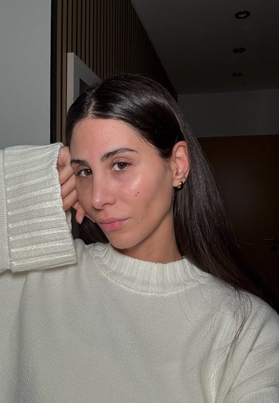 Suéter blanco con textura y cuello de canalé. Cabello largo y oscuro. Pendientes de aro dorados. Tono de piel natural con maquillaje mínimo. Entorno interior.