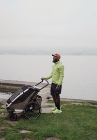 Barnevogn med svart og beige ramme, store hjul og nettingpaneler. Brukeren har på seg en lysegrønn jakke og oransje lue, og står på gress nær vannet.