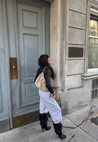 Vestindo uma jaqueta preta e calças de moletom cinza, uma pessoa passa por uma porta cinza texturizada, carregando uma bolsa bege e usando botas de salto alto pretas.