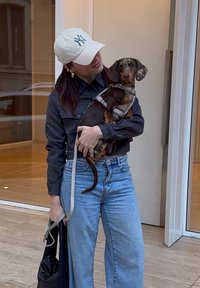 Camicetta a righe nere, jeans azzurri chiari, cappello crema con 'NY', bassotto marrone e beige con imbracatura, borsa nera, in piedi all'aperto.