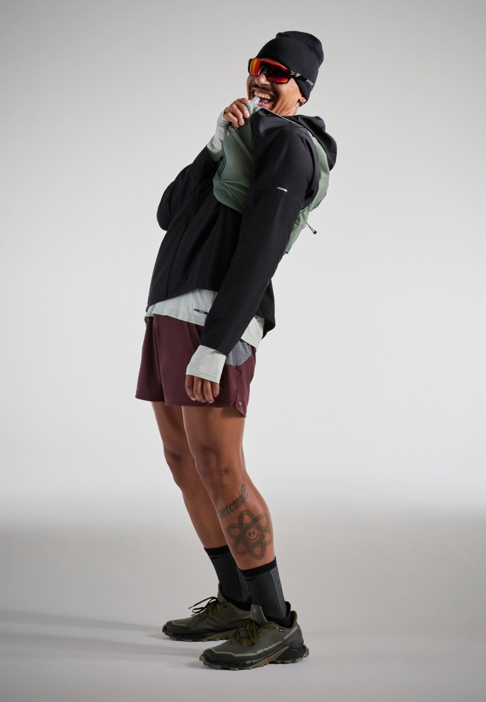 Ľahká čierna bunda so zelenou vestou, vínové šortky a šedé tenisky. Obsahuje čiapku a slnečné okuliare; celkový atletický dizajn.