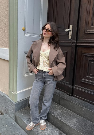 Abrigo de lana marrón sobre una camiseta amarilla con flores, jeans rectos grises y zapatillas grises. Los accesorios incluyen gafas de sol y un bolso pequeño.