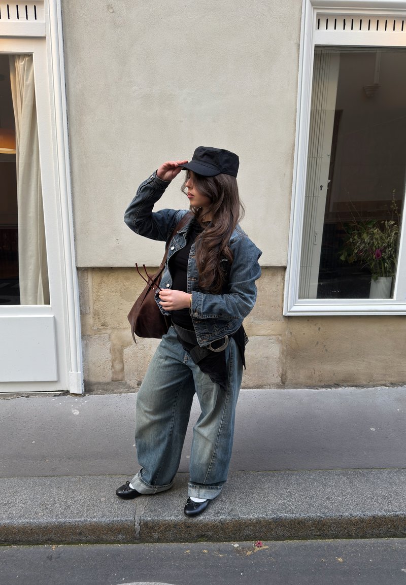 Young woman in denim jacket and jeans adjusts black cap while standing on a narrow urban sidewalk next to beige building with windows.