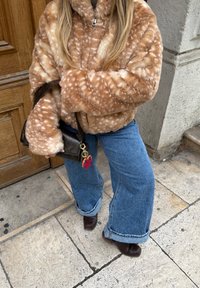 Veste en fausse fourrure beige avec des taches blanches, jeans larges bleus, bottines marron et un sac marron avec un porte-clés en accessoire.