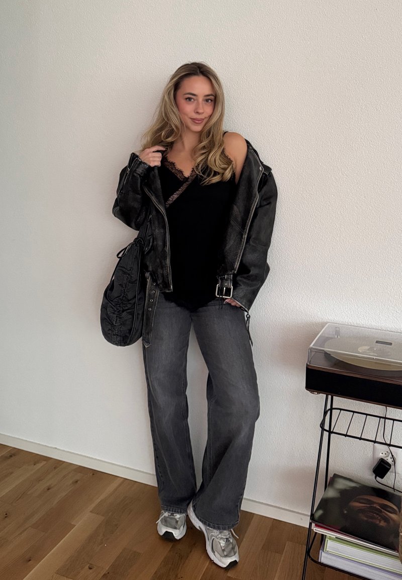 Veste en cuir noire portée sur un haut noir orné de dentelle, jean gris à jambes larges et baskets argentées. Sac noir et tourne-disque à proximité.