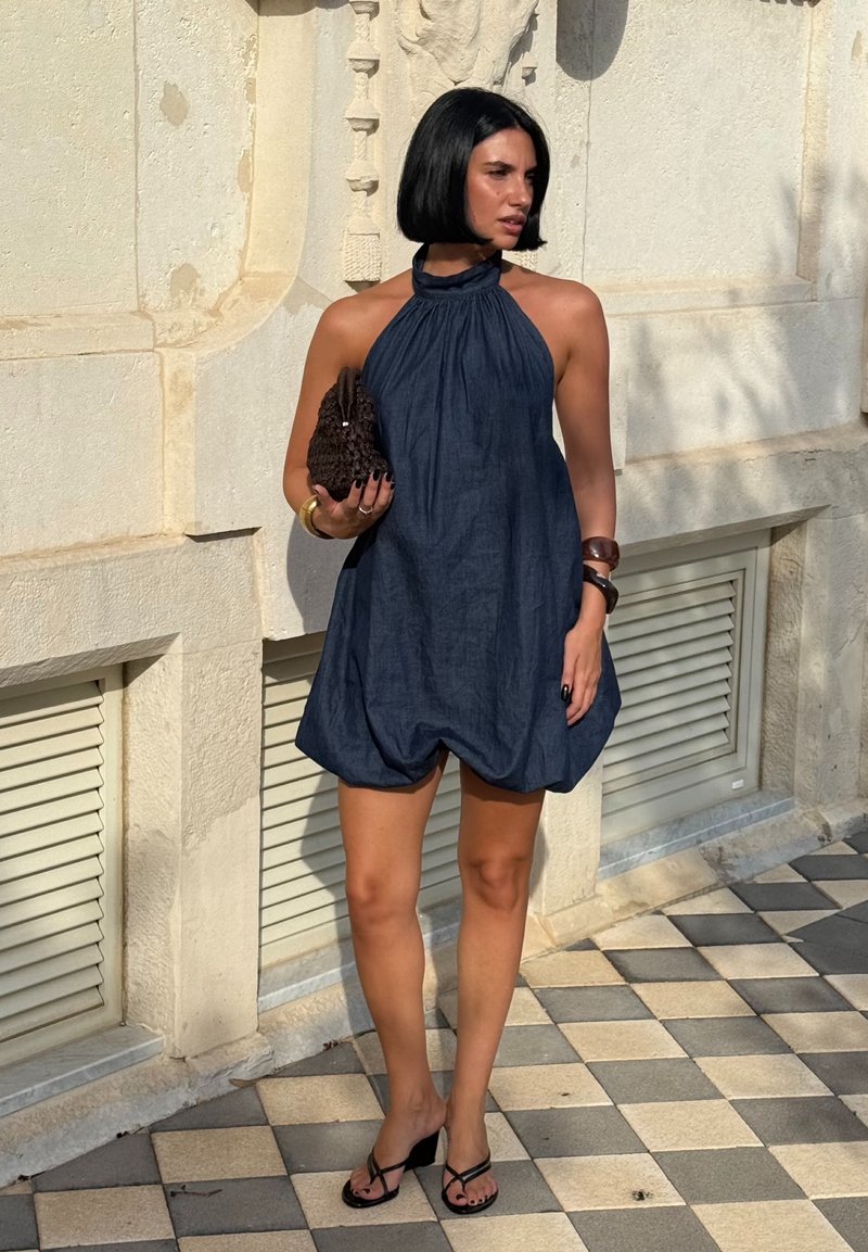 Donna con capelli neri corti che indossa un vestito senza maniche con orlo a bolla blu navy, mentre tiene una clutch marrone con texture, in piedi su pavimentazione piastrellata.