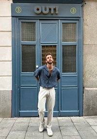 Camisa azul texturizada, calças beje claras e ténis brancos em frente a uma porta azul com painéis de rede castanha e um sinal de "FORA!".