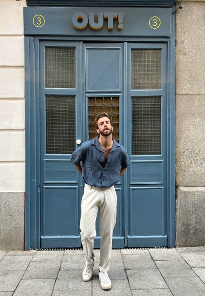Camisa de botones texturizada azul, pantalones beis claros y zapatillas blancas frente a una puerta azul con paneles de malla marrón y un cartel que dice "¡SALIDA!".