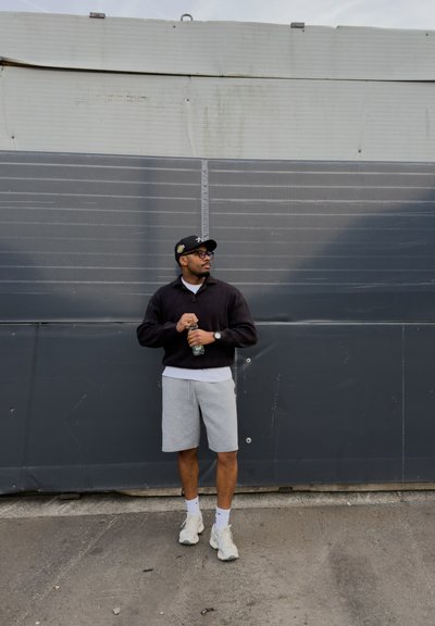Hombre con gorra negra, sudadera negra, pantalones cortos grises y zapatillas blancas, de pie sosteniendo una botella de agua frente a una pared industrial gris.