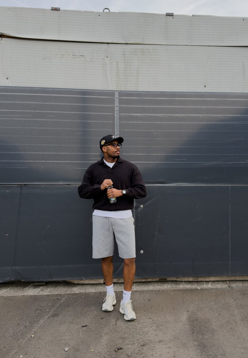 Homme portant une casquette noire, un sweat-shirt noir, un short gris et des baskets blanches, tenant une bouteille d'eau devant un mur industriel gris.