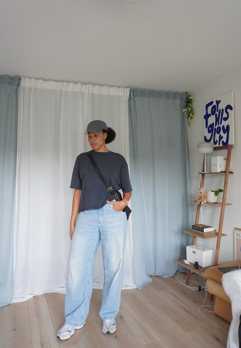 T-Shirt oversize grigio scuro, jeans a gamba larga blu chiaro, berretto grigio e sneakers bianche. Borsa a tracolla nera, mensola in legno e tende sullo sfondo.