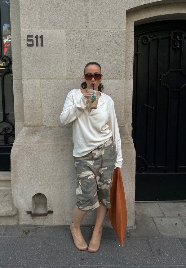 Femeie cu ochelari de soare, pulover alb, pantaloni scurți de camuflaj și pantofi cu toc se sprijină de o clădire, bea cafea cu gheață și ține o geantă mare maro.