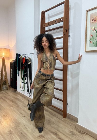 Top corto marrón sin mangas con detalle de cordones, combinado con pantalones marrones holgados. Botas negras. Barras de gimnacio de madera en el fondo. Decoración neutra.