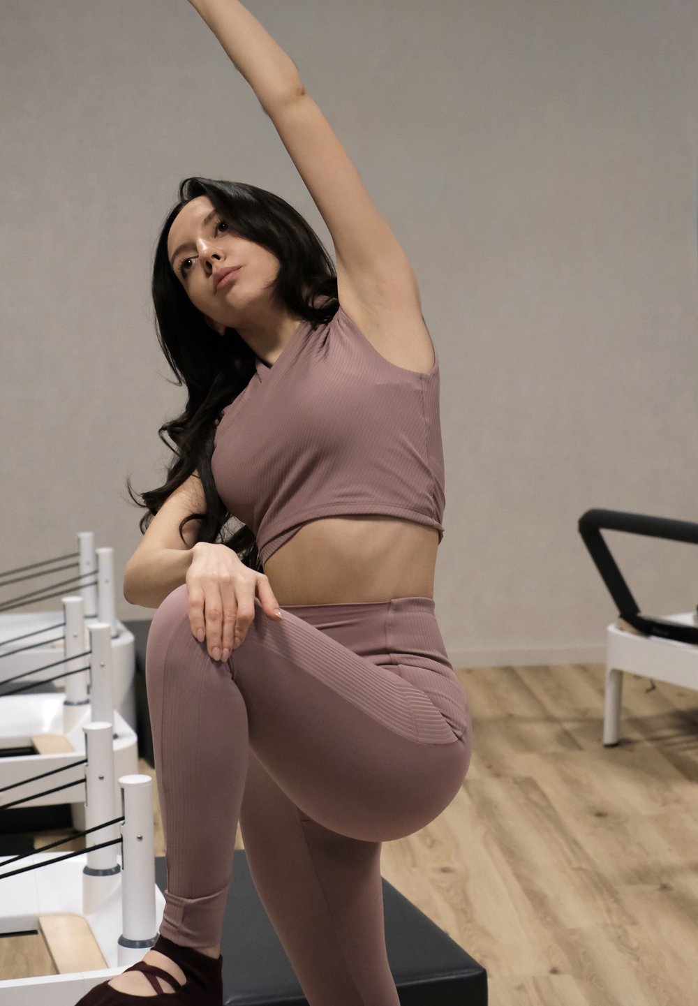 Femeie într-un costum de antrenament mov deschis la culoare, întinzând un braț în sus în timp ce ține genunchiul opus, într-un studio de Pilates cu aparate reformer.