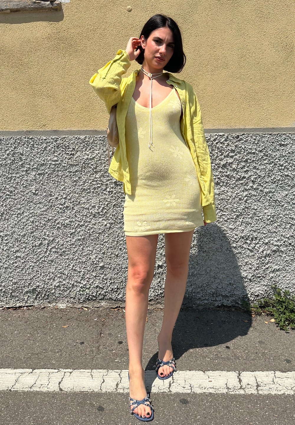 Żółta sukienka z prążkowanego materiału w kwiatowy wzór, nałożona na lekką, otwartą żółtą kurtkę. Ciemnoniebieskie sandały z akcentami z koralików. Neutralne tło.