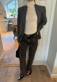 Blazer gris sobre un suéter de cuello alto color crema, combinado con jeans negros lavado ácido con aberturas laterales y zapatos con patrón blanco y negro.