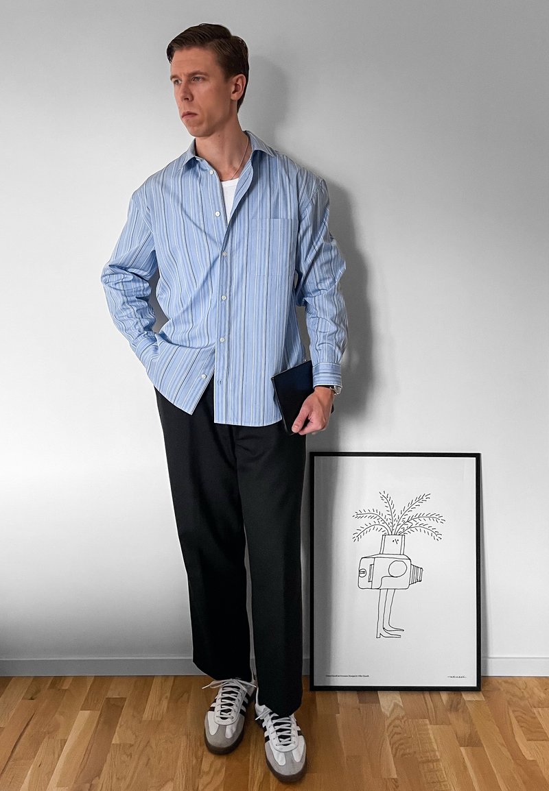 Blue and white striped button-up shirt, black trousers, and grey trainers. Holding a dark notebook; minimalist art print in the background.