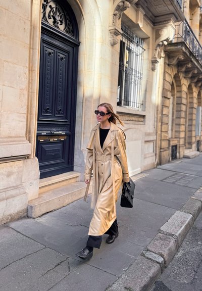 Mujer con gabardina beige y gafas de sol negras caminando por una acera urbana junto a edificios de piedra ornamentados con una gran puerta negra.