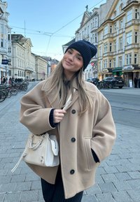 Beige uldfrakke med oversized pasform, sorte knapper og sidelommer. Sort strikket hue og lys beige håndtaske med flettet håndtag.