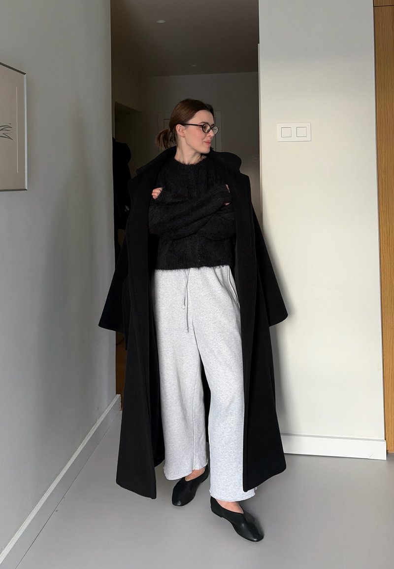 Femme portant un long manteau noir par-dessus un pull noir, un pantalon de survêtement gris clair, des chaussures noires faciles à enfiler et des lunettes, debout à l'intérieur les bras croisés.