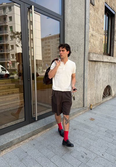Joven caminando por la acera de la ciudad con camisa blanca, pantalones cortos negros, calcetines rojos, zapatos negros, lleva una mochila y sostiene las llaves.
