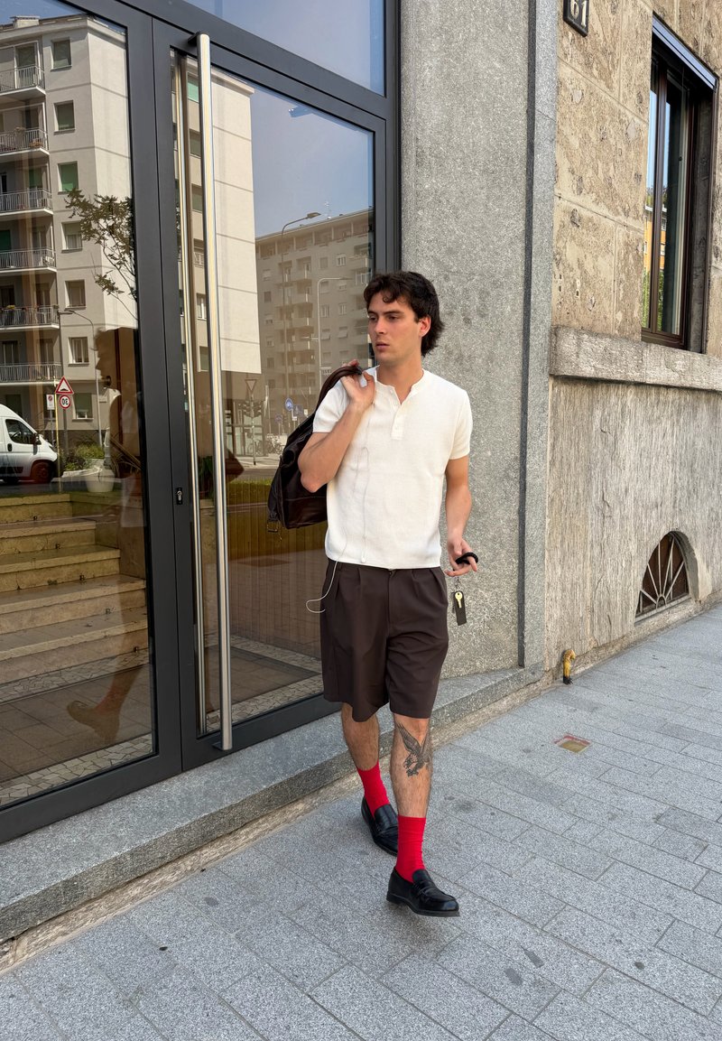Jeune homme marchant sur le trottoir en ville, portant une chemise blanche, un short noir, des chaussettes rouges, des chaussures noires, portant un sac à dos et tenant des clés.