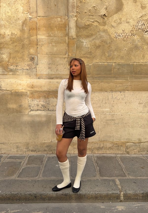 Jeune femme debout sur le trottoir contre un mur en pierre beige texturé, portant un haut blanc à manches longues, un short noir, des chaussettes montantes et tenant une boisson.