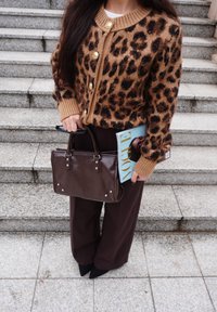 Cardigan con stampa leopardata marrone e nera, bottoni dorati, abbinato a pantaloni marroni a gamba larga e borsa in pelle marrone. Tiene in mano una rivista.
