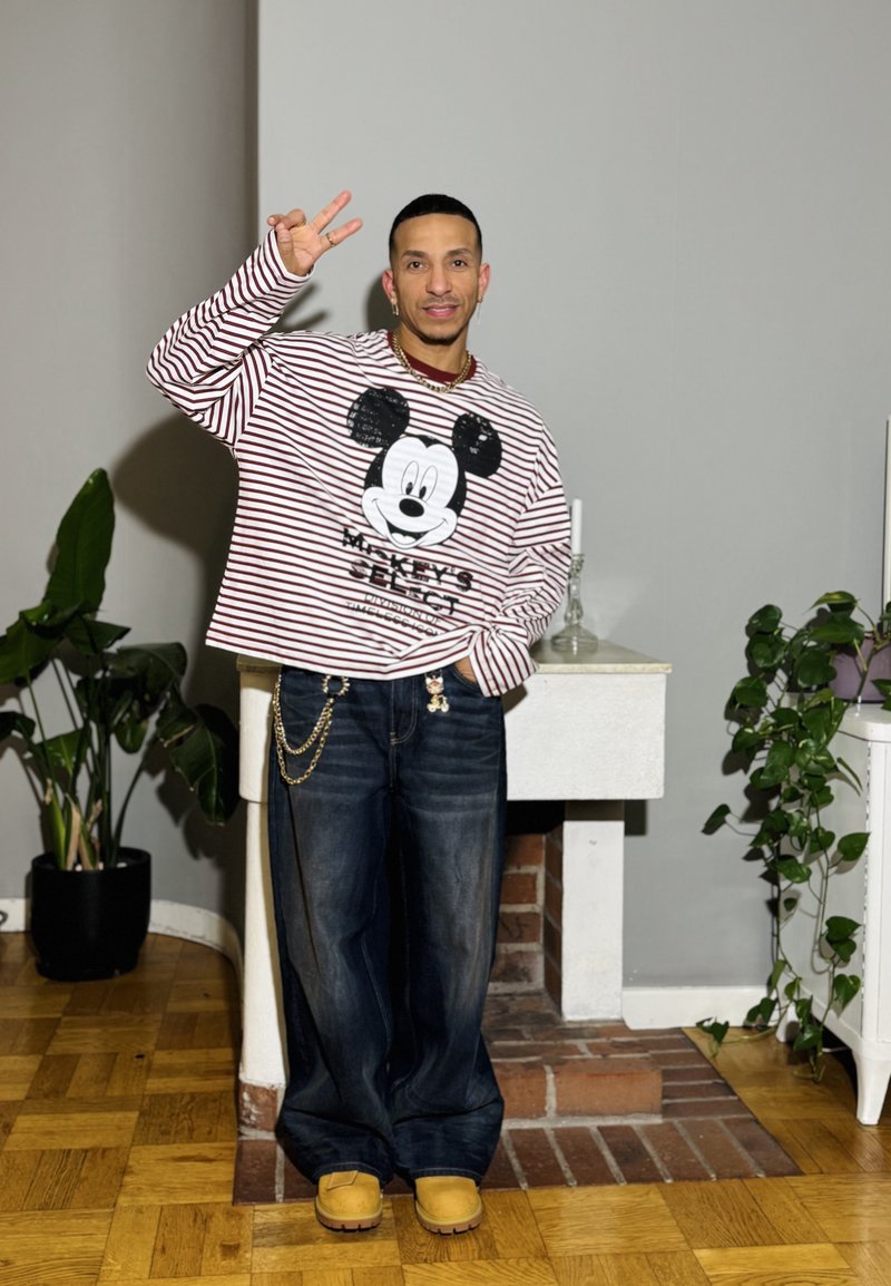 Camisola de manga comprida riscada com gráfico do Mickey Mouse, jeans escuros de ganga e tamancos castanhos. Joalharia simples e plantas interiores ao fundo.