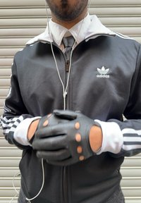 Άνδρας με μαύρο μπουφάν Adidas, λευκό πουκάμισο, γραβάτα, μαύρα δερμάτινα γάντια χωρίς δάχτυλα και λευκά ακουστικά, στέκεται με τα χέρια ενωμένα.