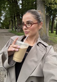 Beige trenchcoat; mörk topp med utskärningsdesign; bruna glasögon; håller en klar kopp med iskallt kaffe; utomhusmiljö med träd.