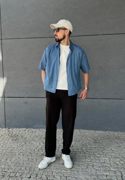 Camisa de botones de manga corta azul clara sobre una camiseta blanca, pantalones negros y zapatillas blancas, con una gorra beige y gafas de sol.