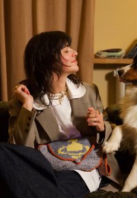 Femme en blazer à carreaux assise sur un canapé, interagissant avec un chien qui lève la patte, tenant un sac tricoté gris et orange sur ses genoux.