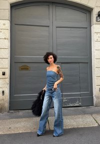 Kvinna med lockigt hår och tatuering på armen, iförd denimtop och vida jeans, står framför en stor grå träport och håller en svart väska.