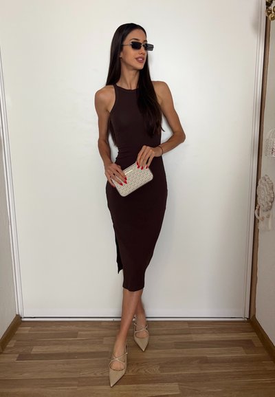 Mujer con vestido marrón oscuro sin mangas que sostiene un bolso de mano beige, lleva gafas de sol y zapatos de punta beige, de pie sobre un suelo de madera.