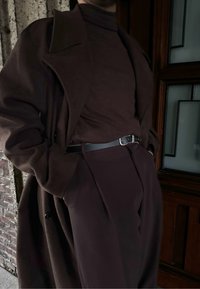 Σκούρο καφέ oversized παλτό από μαλακό ύφασμα, φορεμένο πάνω από σκούρο φούτερ με ζιβάγκο. Διαθέτει μια λεπτή μαύρη ζώνη. Το υπόβαθρο παρουσιάζει ξυλώδεις και τούβλινες υφές.