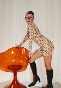 Γυναίκα σε μπεζ κορμάκι με ημισεληνοειδή μοτίβα, μεγάλα γυαλιά και μαύρα μποτάκια με κορδόνια, στηρίζεται σε διαφανή πορτοκαλί καρέκλα μπροστά από λευκή κουρτίνα.