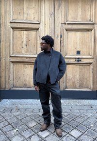 Veste en laine gris foncé avec col montant et fermeture à boutons, associée à un jean noir et des bottines marron, sur un fond en bois.