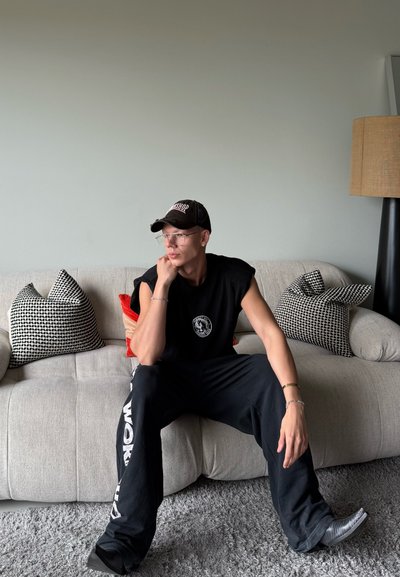 Gorra negra con logo, gafas claras, camiseta sin mangas negra, pantalones negros con texto blanco, sentado en un sofá gris claro con cojines estampados.