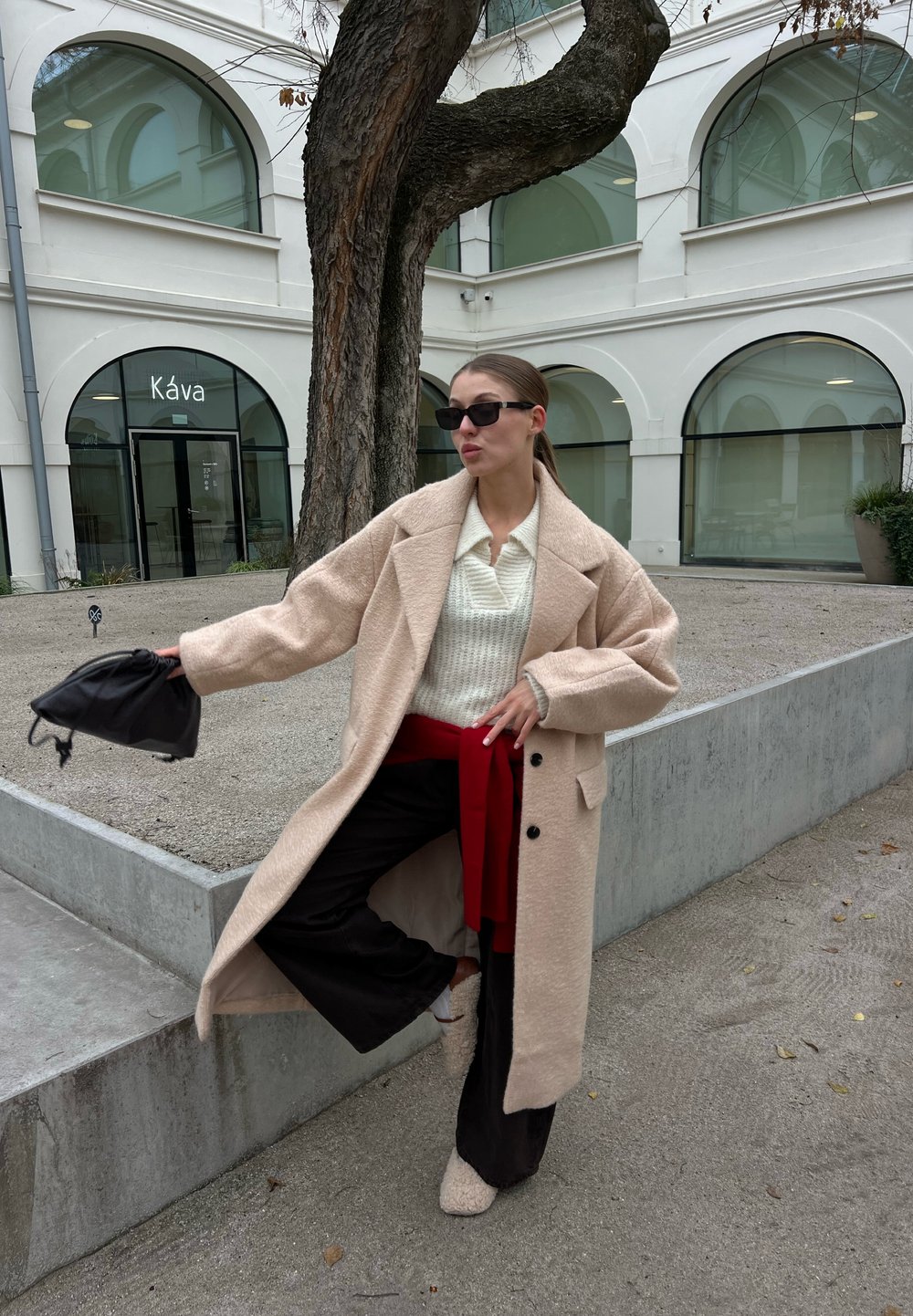 Femme en manteau beige, pull blanc, pantalon noir et lunettes de soleil, tenant un sac noir, posant près d'un arbre et d'un bâtiment blanc avec des fenêtres en arc.